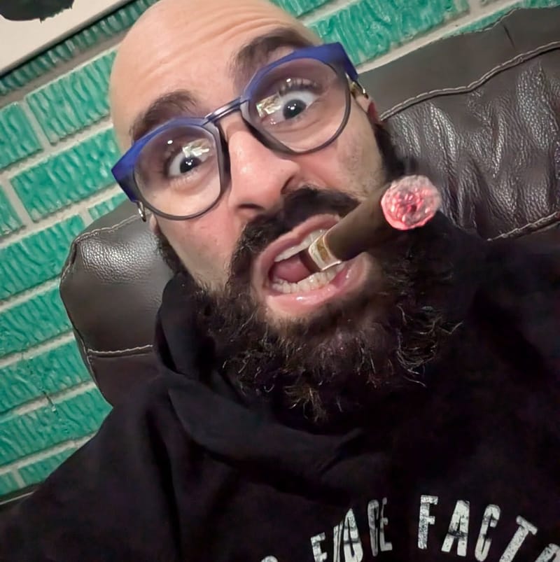 Very close selfie against a green brick wall, cigar clamped in his teeth, eyes wide behind heavy glasses.