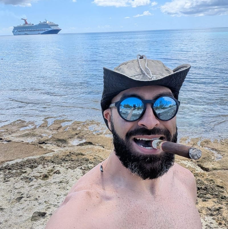 Abe shirtless on a rocky shoreline in a wide sun hat and mirrored sunglasses with a cigar in his teeth, a cruise ship on the horizon behind him.