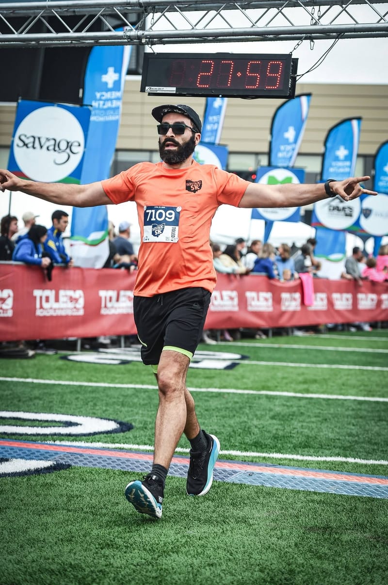 Abe crossing a finish line on a football field with both arms flung wide, bib 7109, the overhead race clock reading 27:59.