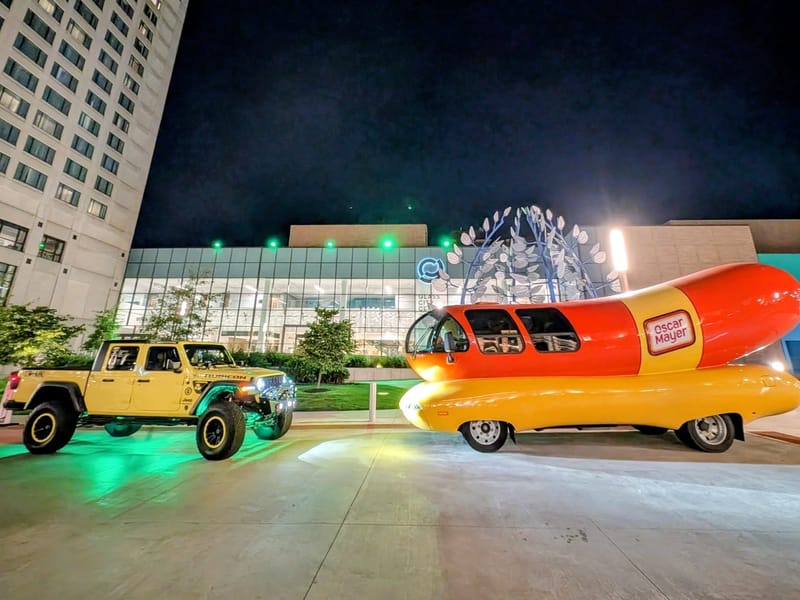The yellow Jeep parked at night beside the Oscar Mayer Wienermobile, both lit from below.