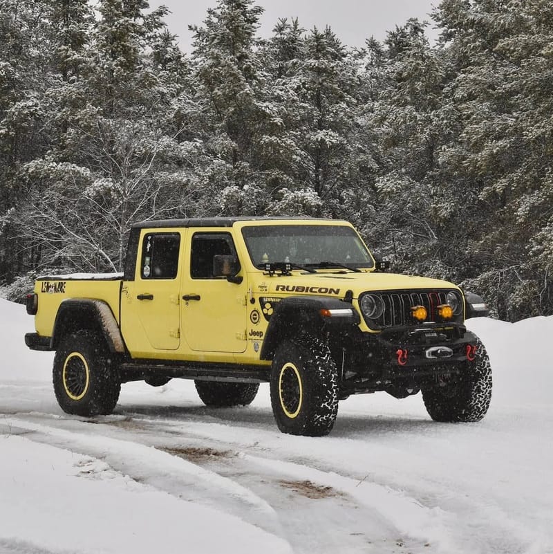 The yellow Jeep Gladiator Rubicon parked in deep snow with snow-loaded evergreens behind it.