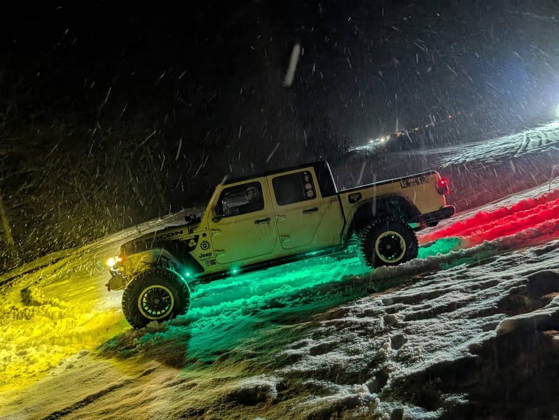 The yellow Jeep at night in falling snow, yellow and green underglow throwing color across the ground.