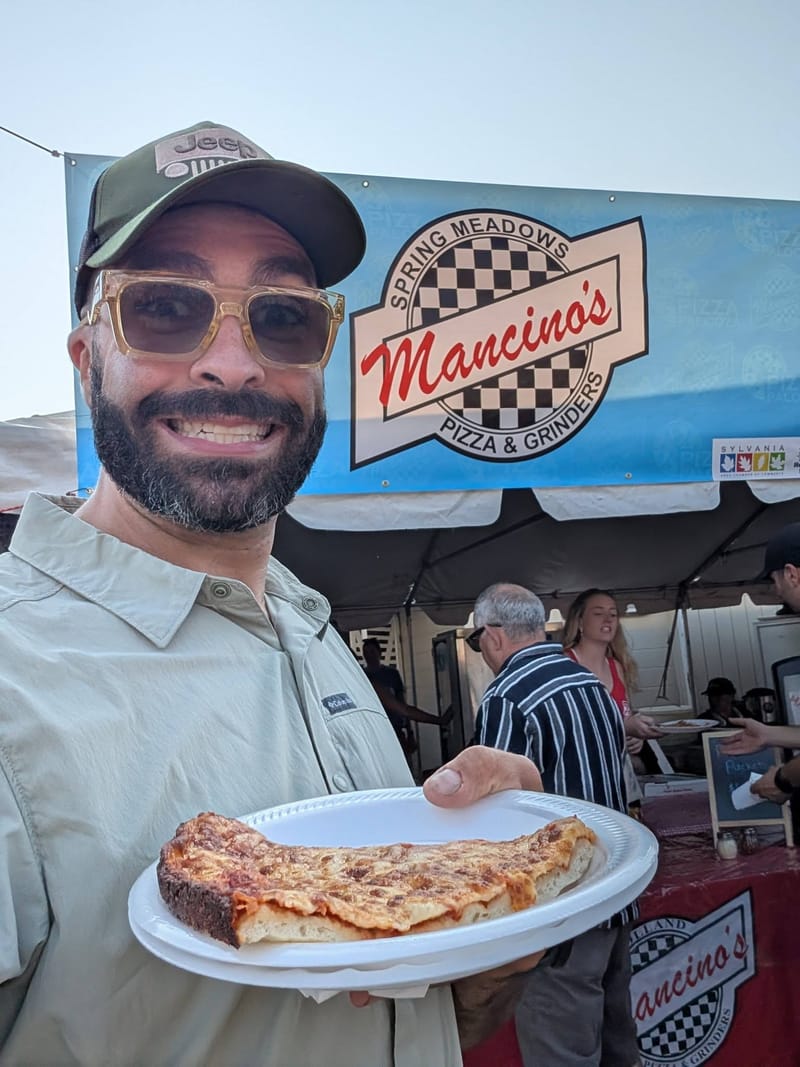 Abe holding a paper plate with a large slice of pizza in front of a Mancinos festival stand.