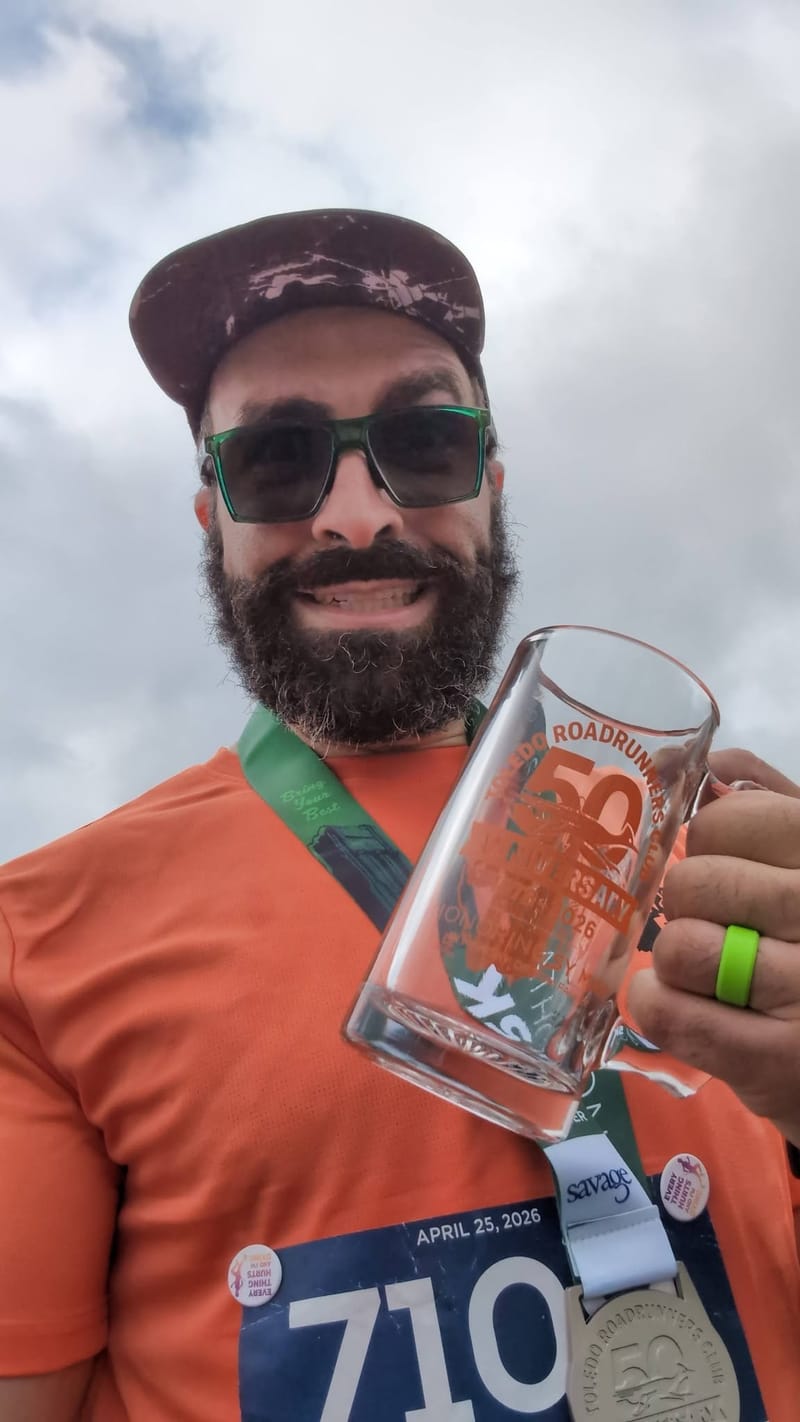 Post-race selfie under grey sky, finisher medal round his neck and a commemorative glass mug held up, bib dated April 25 2026.