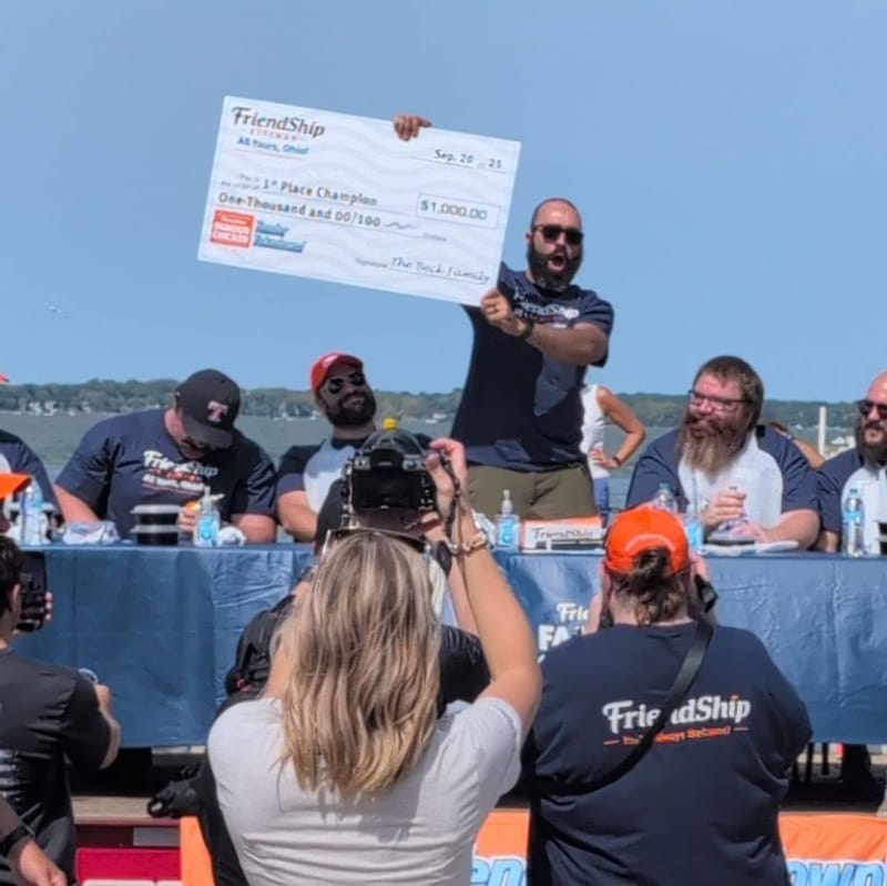 Abe on a riverside stage holding the winning check overhead while a crowd and camera crew watch from below.