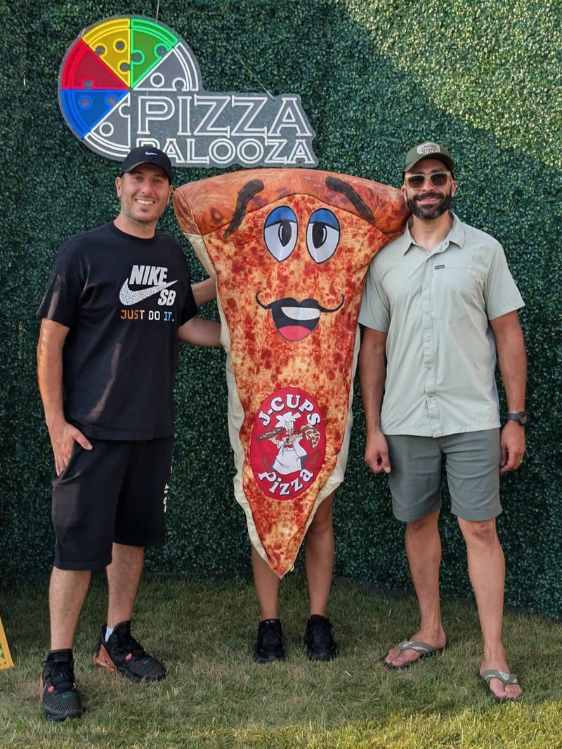 Three men standing either side of a giant cartoon pizza-slice mascot at a Pizza Palooza backdrop.